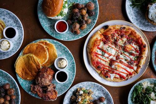 A spread of various dishes including pancakes, fried chicken, pizza, and small bites on decorative plates.