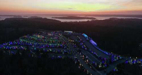 Aerial view of a colorful holiday light display with trees and decorations, set against a sunset over a lake.