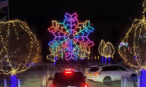 Colorful illuminated snowflake display surrounded by trees adorned with lights at night. Cars are parked nearby.