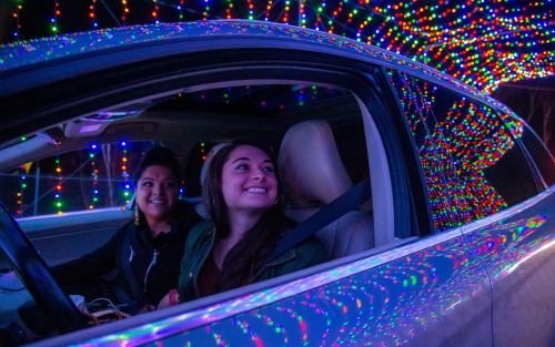 Two women smile inside a car, surrounded by colorful holiday lights illuminating the scene.