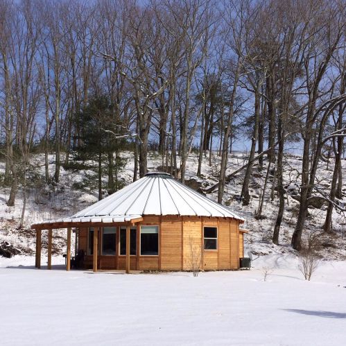 A wooden yurt with a circular roof sits in a snowy landscape, surrounded by bare trees under a clear blue sky.