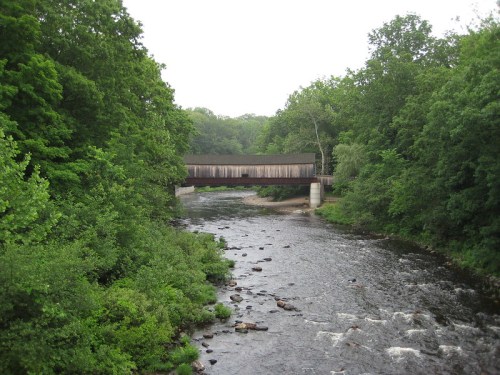A wooden covered bridge spans a river, surrounded by lush green trees and rocky riverbanks.
