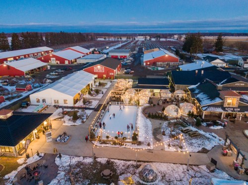 Aerial view of a winter market with an ice skating rink, festive lights, and surrounding red barns at dusk.