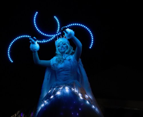 A performer in a glowing blue costume and crown twirls illuminated props against a dark background.