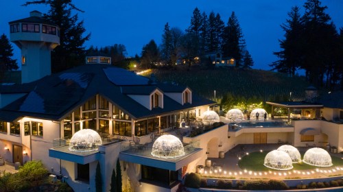 A scenic view of a winery at night, featuring illuminated dining igloos and a modern building surrounded by trees.