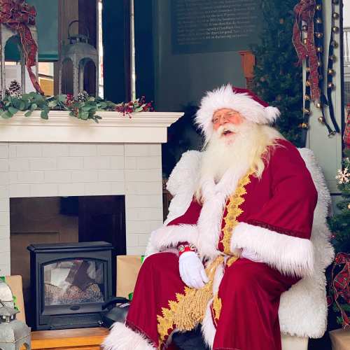 A jolly Santa Claus in a red suit sits by a cozy fireplace, surrounded by festive decorations.