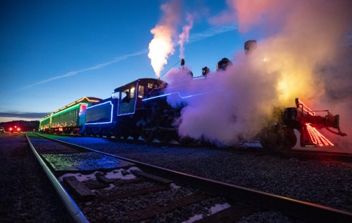 A brightly lit steam train emits smoke at dusk, with colorful lights adorning its cars along the tracks.