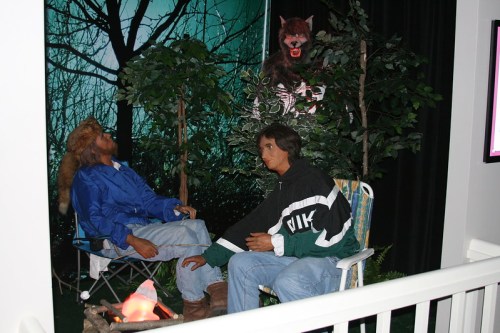 Two life-like figures sit by a campfire in a forest setting, with a wolf figure looming above them in the background.