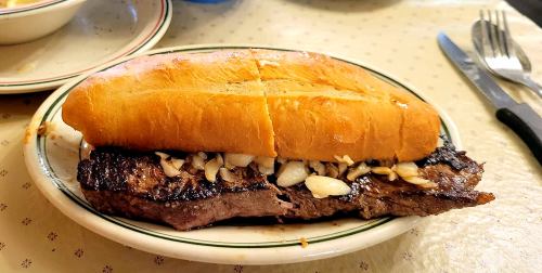 A grilled steak sandwich with sliced onions on a toasted bun, served on a plate with utensils in the background.