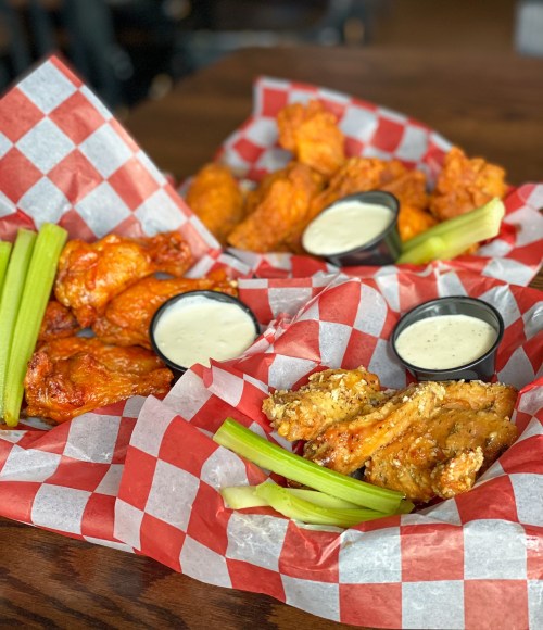 Two baskets of chicken wings with celery sticks and dipping sauces, served on red and white checkered paper.
