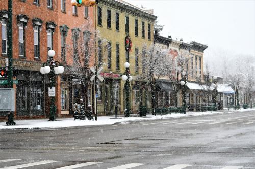 Snow falls gently on a quaint street lined with colorful buildings and festive decorations, creating a winter wonderland.