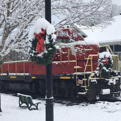 A red train engine decorated with holiday wreaths, surrounded by snow and a festive atmosphere.