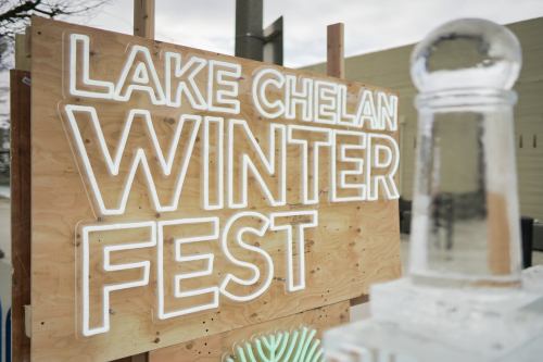Sign for Lake Chelan Winter Fest, featuring bright white letters on a wooden background, with an ice sculpture in the foreground.