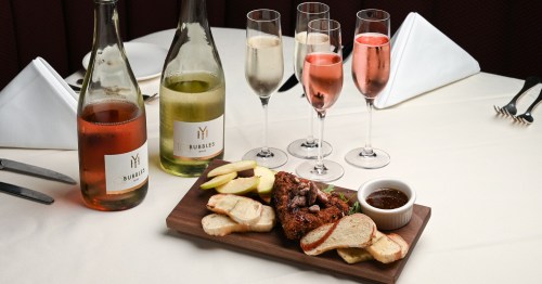 A wooden platter with fried chicken, bread, apple slices, and dipping sauce, alongside bottles of wine and champagne flutes.