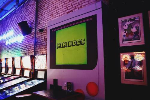 A retro arcade wall featuring a large "MINIBOSS" screen and pinball machines in a vibrant, colorful setting.