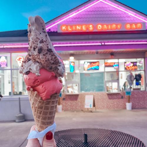 A hand holds a colorful ice cream cone in front of Kline's Dairy Bar, illuminated with neon lights at dusk.