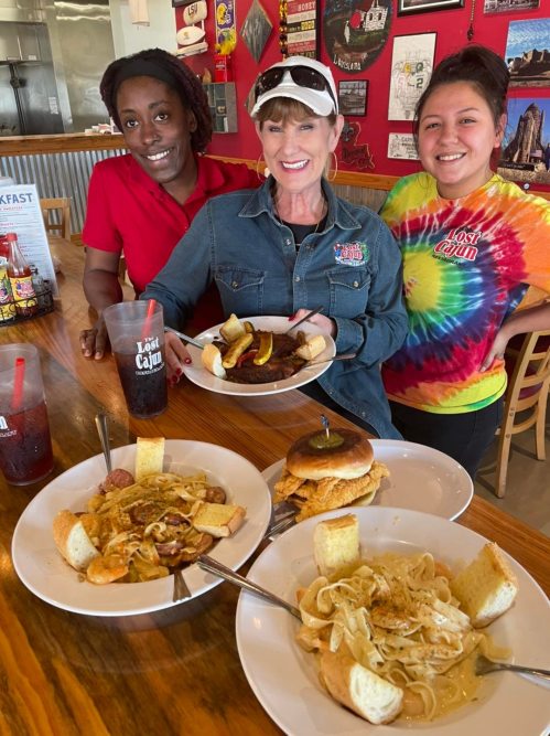 Three women smile in a restaurant, holding plates of food, with dishes of pasta and sandwiches on the table in front of them.