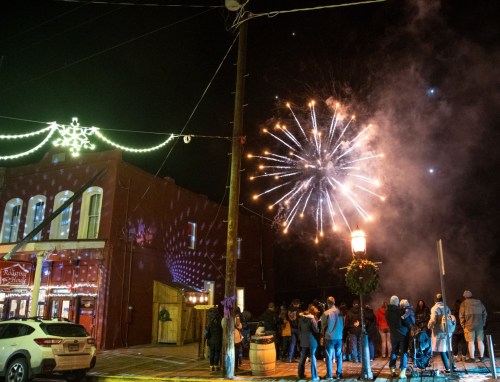 A crowd watches fireworks illuminate the night sky, with festive lights and decorations on nearby buildings.