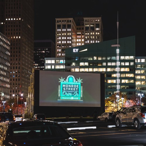 A nighttime view of a drive-in movie screen displaying "Monroe Street Drive-In," surrounded by city buildings and parked cars.