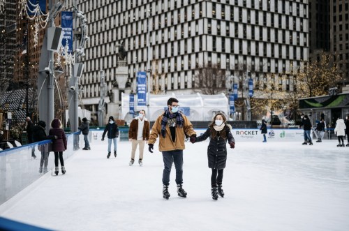 A couple skates hand-in-hand on an ice rink, surrounded by other skaters in a city setting.