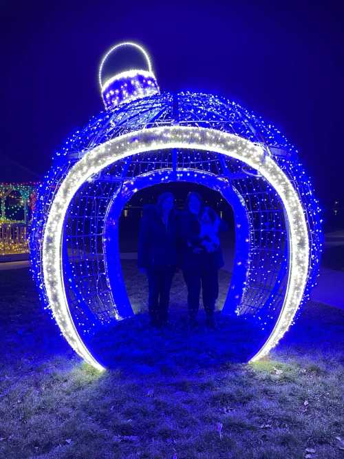 Two people stand inside a large, illuminated ornament structure decorated with blue and white lights at night.