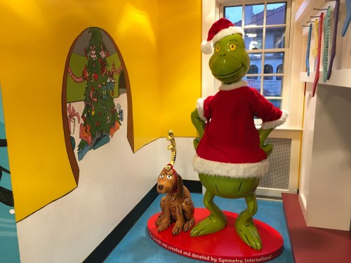 A large Grinch statue in a Santa outfit stands next to a dog, with colorful walls and a Christmas tree mural in the background.