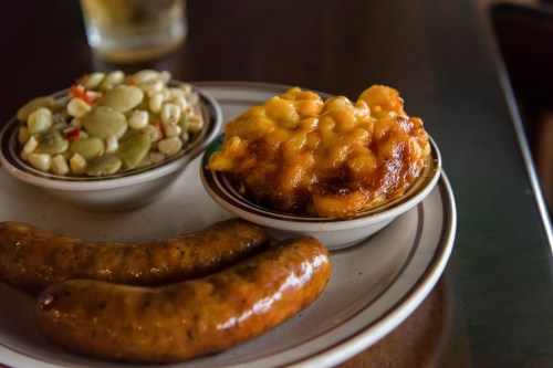 A plate with two sausages, a bowl of corn salad, and a bowl of cheesy macaroni.