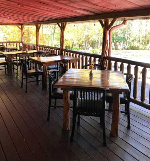 A rustic wooden porch with several tables and chairs, surrounded by trees and autumn foliage.