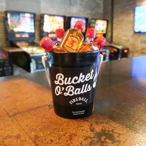 A black bucket filled with mini bottles of Fireball whisky, set against a backdrop of arcade games.