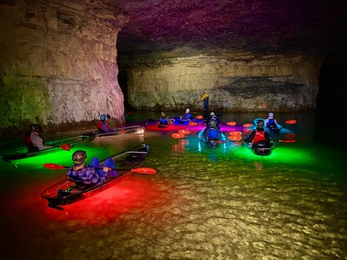 A group of kayakers with colorful lights paddling in a cave, surrounded by illuminated rock formations.