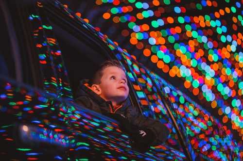 A child gazes out of a car window, surrounded by colorful holiday lights.