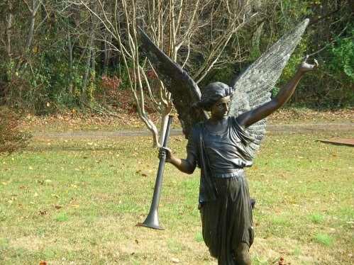 A bronze angel statue with large wings, holding a trumpet, stands in a grassy area surrounded by trees.