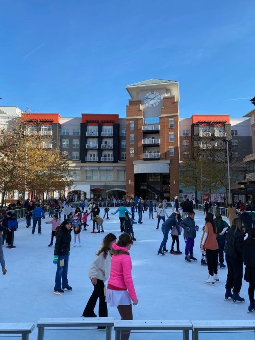 A busy outdoor ice skating rink with people skating, surrounded by buildings and trees under a clear blue sky.