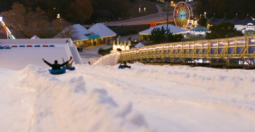 A snowy hill with people tubing down, surrounded by festive lights and a ferris wheel in the background.