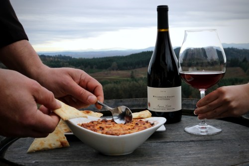 A person serves dip from a bowl with pita, alongside a glass of red wine and a bottle, set against a scenic backdrop.