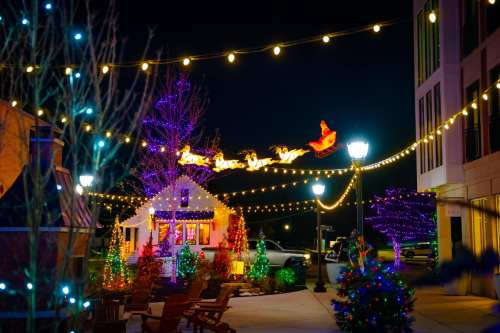 A festive scene with colorful holiday lights, a decorated house, and a sleigh with reindeer in the night sky.