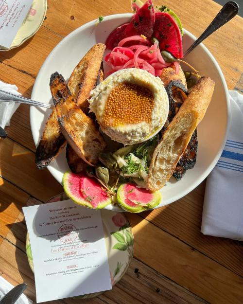 A plate featuring hummus, grilled bread, pickled vegetables, and fresh fruit, set on a wooden table.