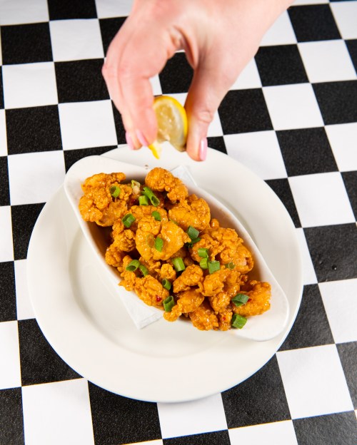 A hand squeezes lemon over a plate of crispy fried chicken pieces garnished with green onions on a checkered tablecloth.