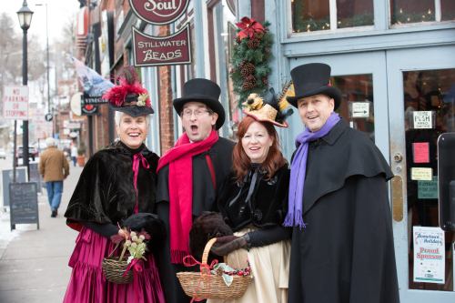 Four carolers in Victorian attire stand outside a shop, smiling and holding baskets, with festive decorations around them.
