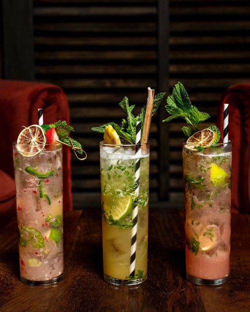 Three colorful cocktails with mint, lemon, and decorative straws, served in tall glasses on a wooden table.