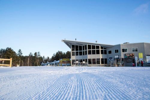 A modern ski lodge with large windows, surrounded by snow-covered ground and trees under a clear blue sky.