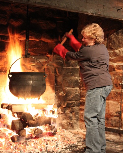 A person stirs a large pot over an open fire in a stone fireplace, wearing red oven mitts. Flames flicker around them.
