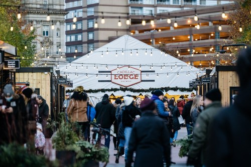 A bustling outdoor market scene with a large tent labeled "Cadillac Lodge" and string lights overhead.