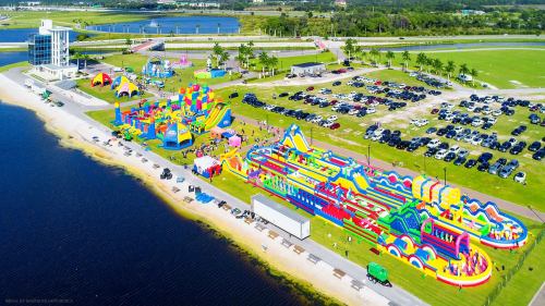Aerial view of a colorful inflatable water park beside a lake, with cars parked nearby and green grass in the background.