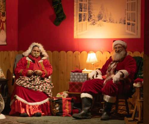 A woman in a festive red dress and Santa Claus sit together in a cozy, decorated room with holiday gifts.