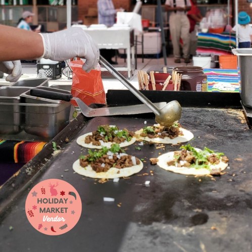 A vendor prepares tacos on a griddle, adding toppings with a spatula, at a holiday market.