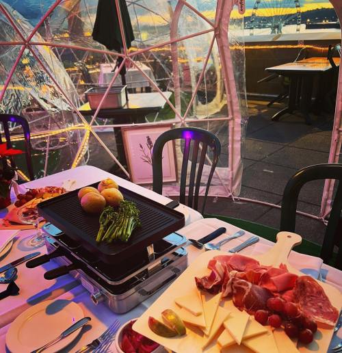 A cozy dining setup inside a transparent dome, featuring a grill, fresh vegetables, and a platter of meats and cheeses.