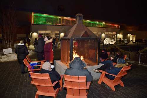 People gather around a fire pit at night, with colorful lights in the background and snow on the ground.