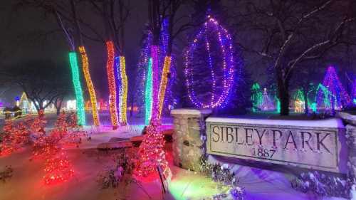 Colorful holiday lights illuminate Sibley Park, with snow-covered trees and festive decorations in a winter night scene.