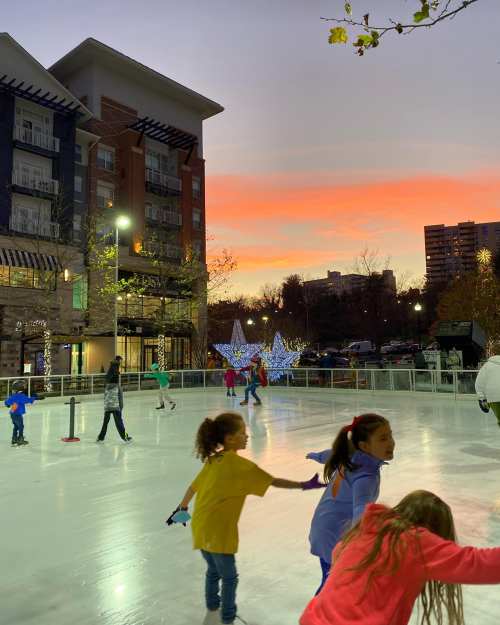 A vibrant ice skating rink at sunset, with people skating and festive lights in the background.